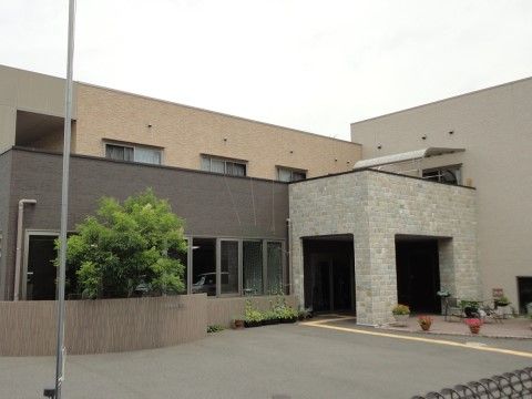 建物はベージュ、茶色、グレーシュで外壁が塗り分けられている。玄関には柱が付いたひさしが設置されていて、その周辺に植木鉢が置かれている。