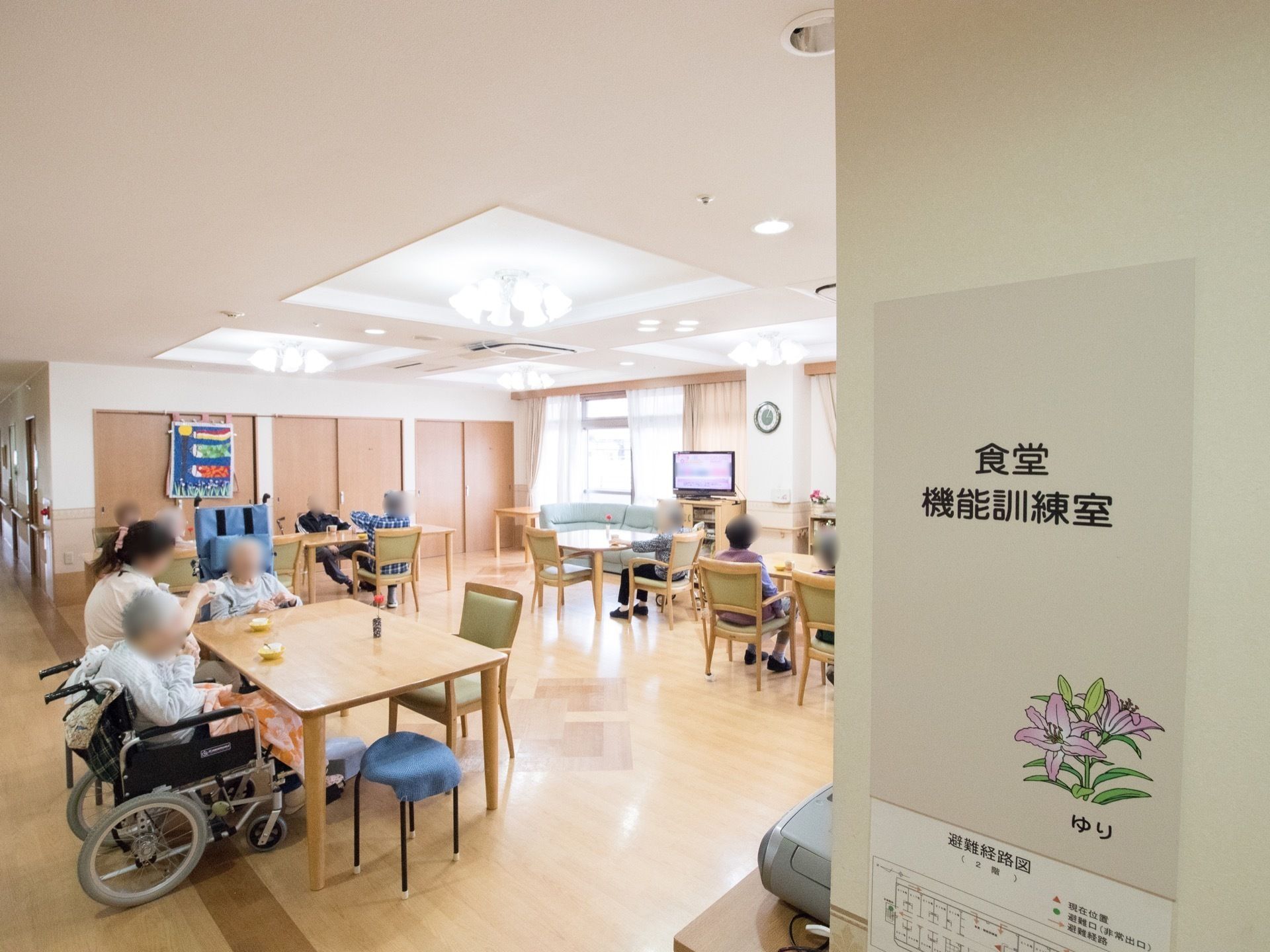 食堂は機能訓練室を兼ねており、壁に案内板の表示がされている。最大４人がけのテーブルセットが用意されている。液晶テレビが置かれている。