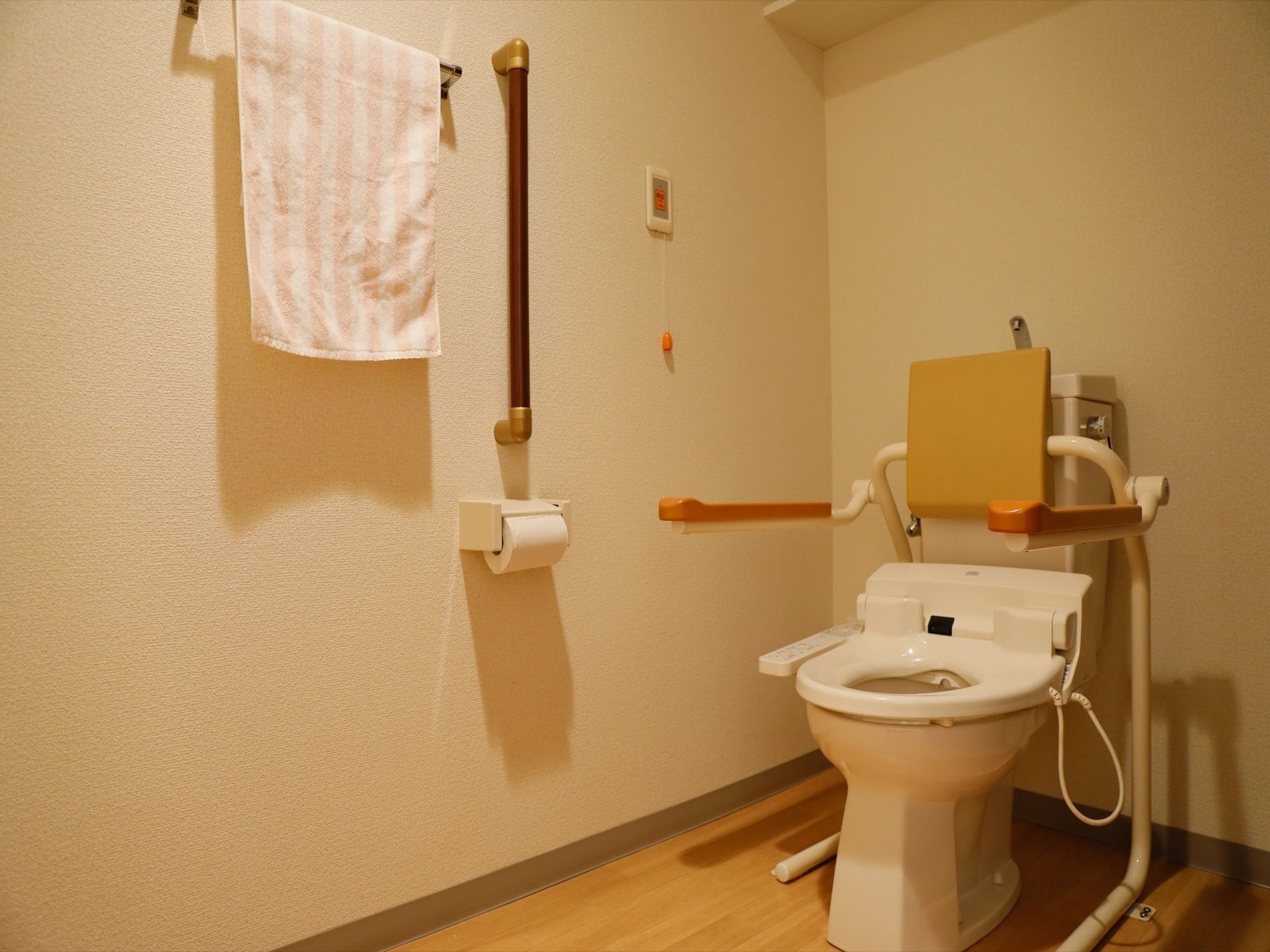 木目調のフローリングを取り入れたトイレである。壁には手すりを備え、隣にタオル掛けを設置している。緊急呼び出しブザーがある。
