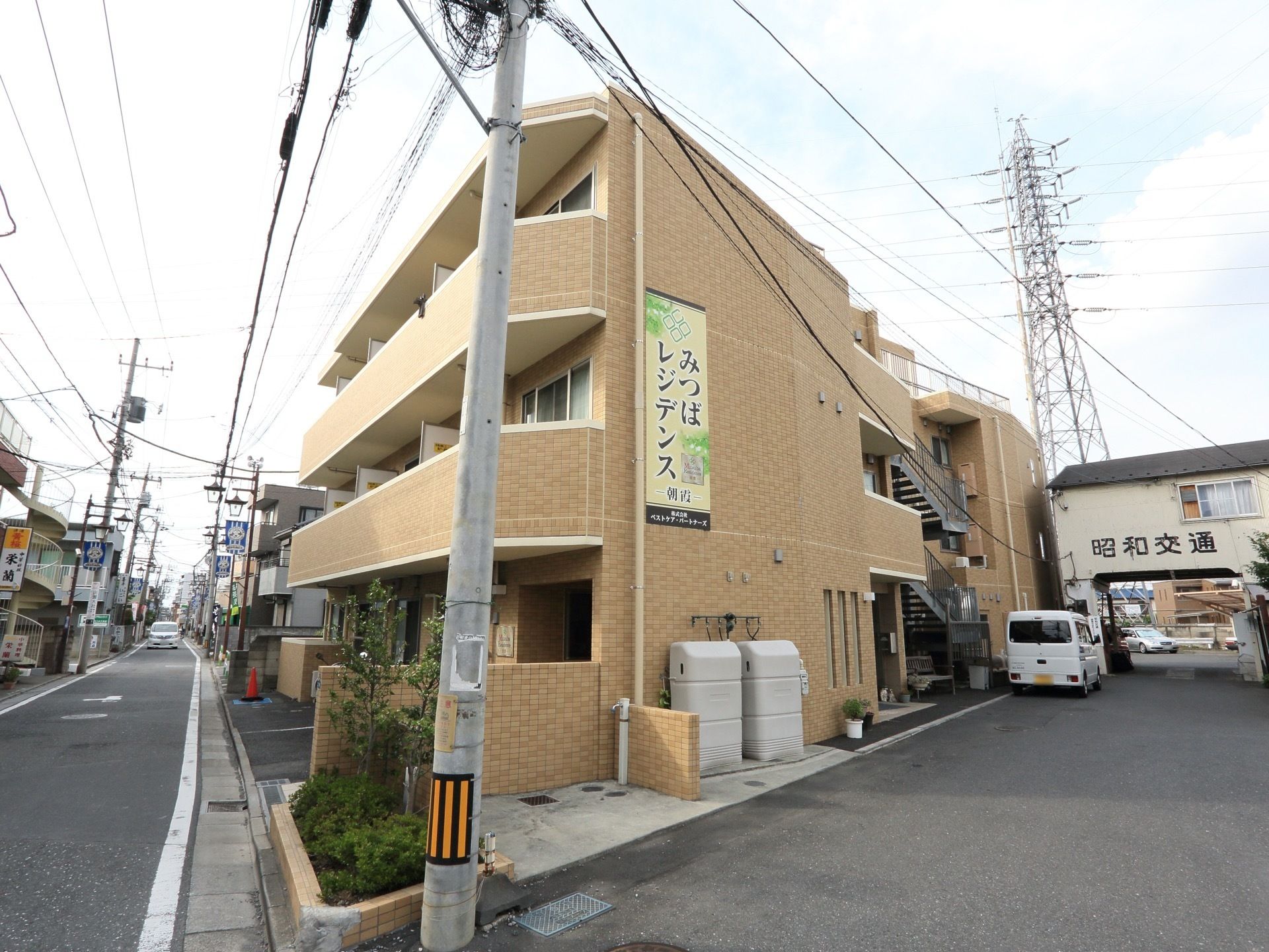 閑静な住宅地に立地する、ブラウン調の建物である。道路に面しており、整備された緑地のとなりには、電柱が立っている。