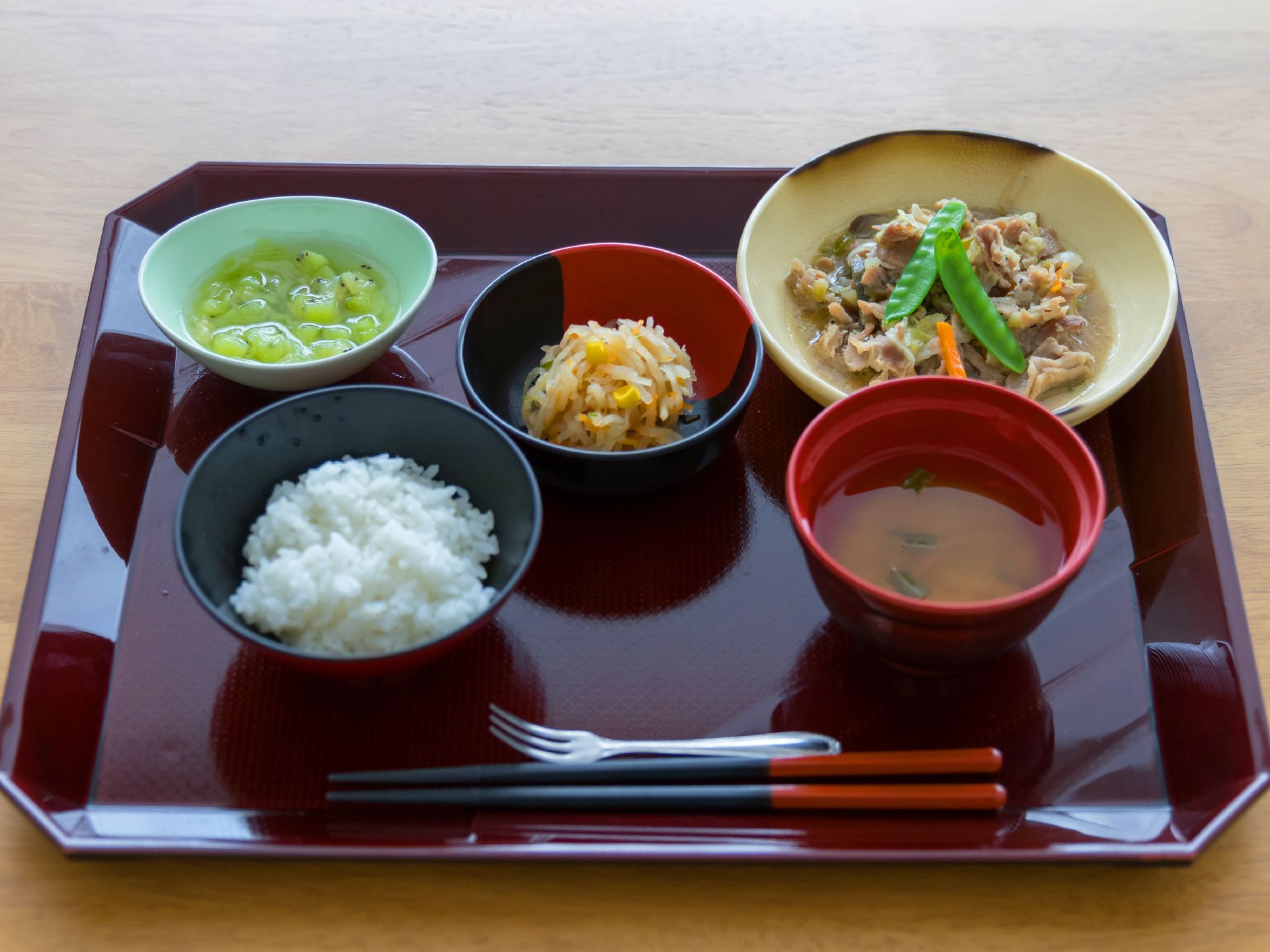 お味噌汁とご飯がお盆の上に乗っている。丸い皿に果物が盛り付けてある。絹さやなどの野菜と肉の炒めが盛り付けてある。