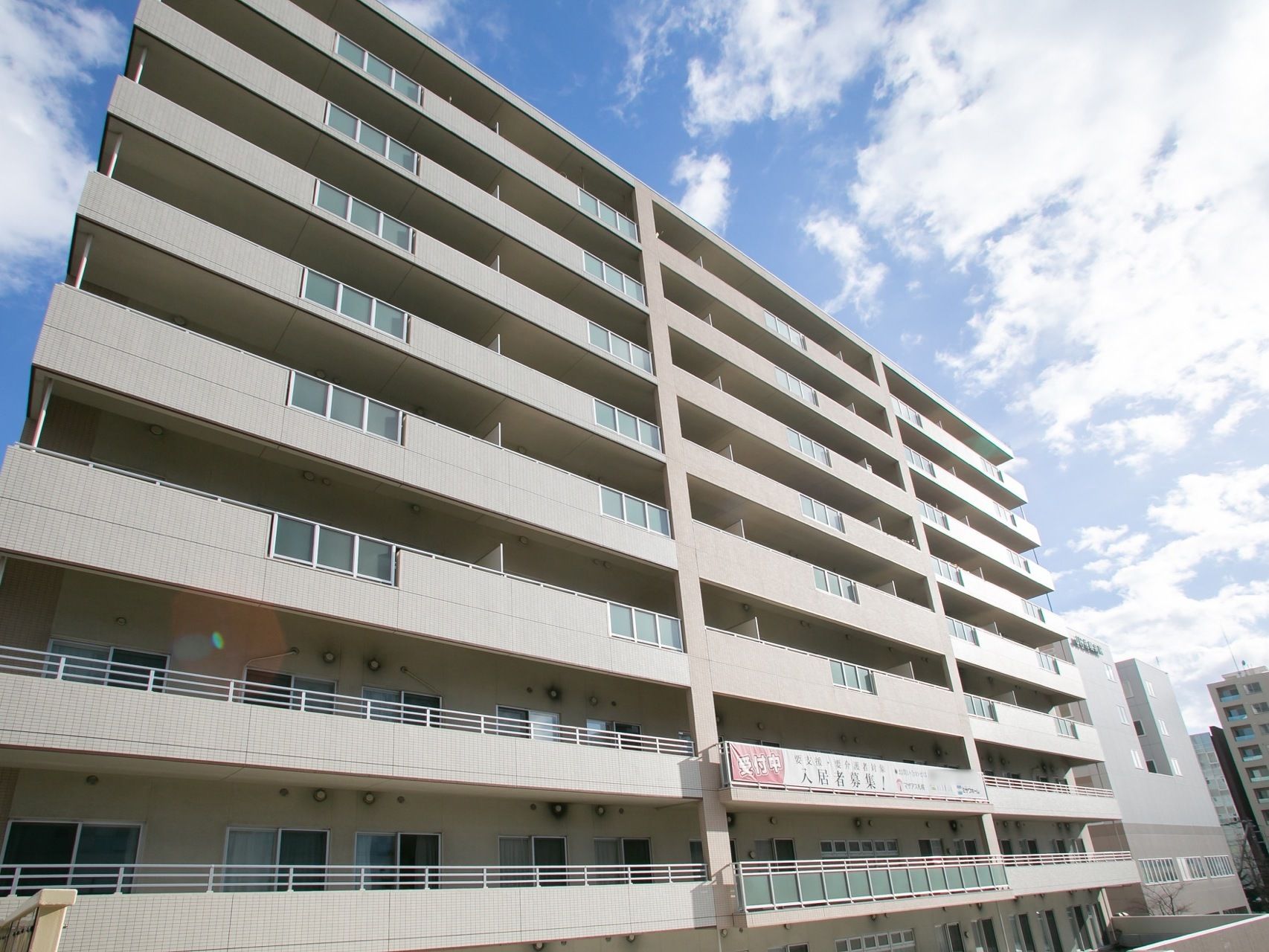 居住フロアが１０階まである施設で、淡い紫色の外観の建物である。低層階に入居者募集の垂れ幕が貼られている。