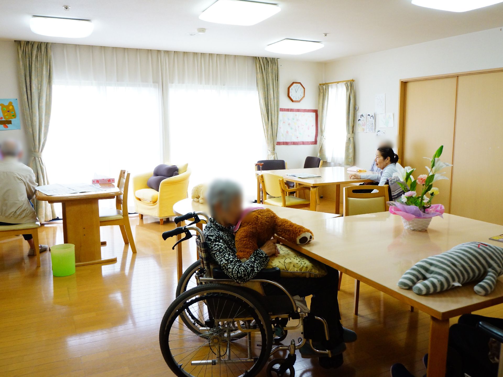 大きな窓のあるリビングである。入居者は思い思いに過ごしている様子がわかる。ぬいぐるみを抱いている女性がいる。