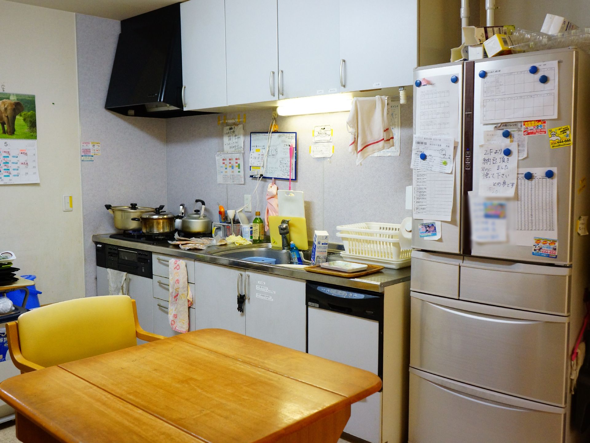 キッチンコンロの上に鍋とやかんが置かれている。冷蔵庫にはたくさんの紙が磁石で貼り付けられている。四角いテーブルと椅子が置いてある、