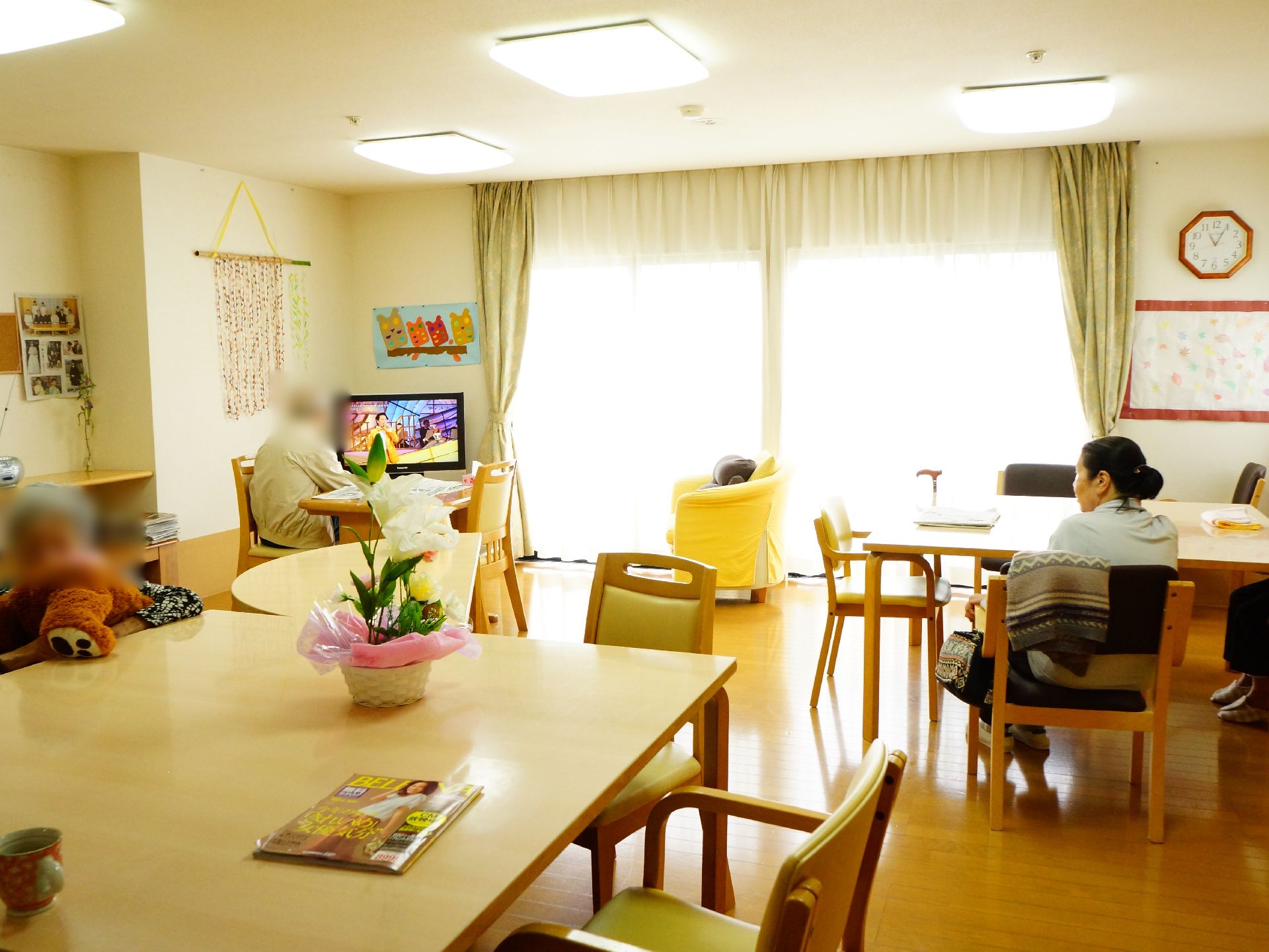 長方形の机の上にはコップと雑誌、お花が置いてある。部屋のテレビを見ている方がいる。壁には写真や絵、時計がある。