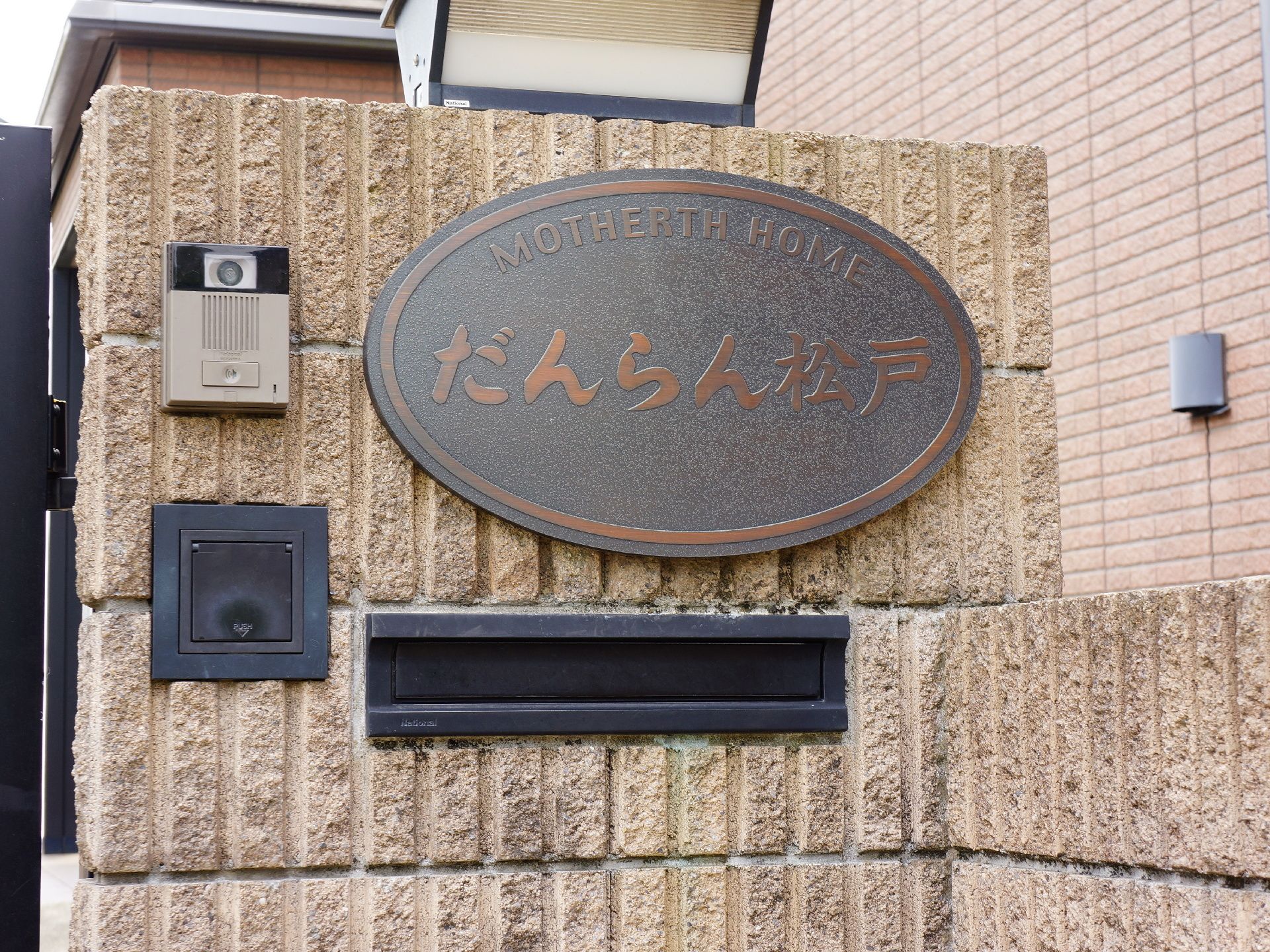 建物外側にはカメラ付きのインターフォンがある。その下にはポストがある。右上には大きくだんらん松戸と書かれた表札がある。