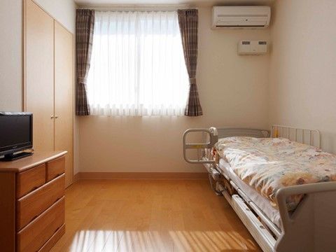 居室にはエアコンが設置され、手すり付きベッドやテレビ、木製のチェストが置かれている。窓があって明るいフローリングルームである。