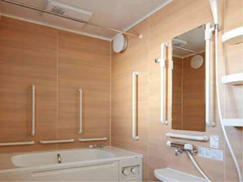 浴室の壁は明るいベージュの木目で、縦型や横型の手すりが多く設置されている。浴槽やシャワー、鏡、カウンターがあり、ゆっくりと入浴を楽しめる。