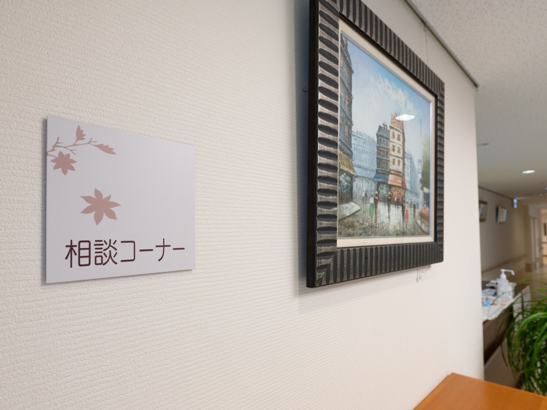 白い壁に相談コーナーと書かれたパネルが貼られている。隣には額に入った絵画が飾られている。観葉植物や棚が置かれている。