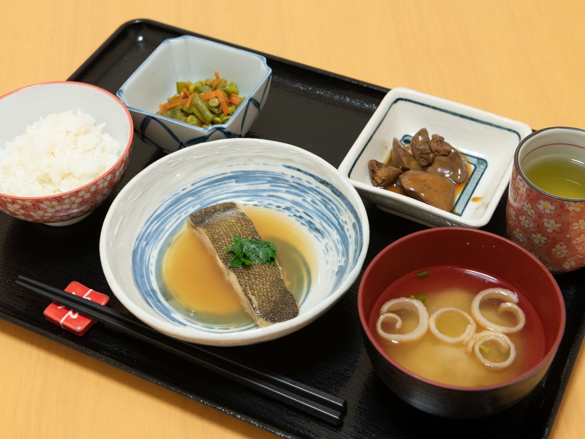 黒いお盆の上に、ごはんとお吸い物、魚の煮つけ、おかずの小鉢が2つ、湯飲みに入ったお茶が置かれている。