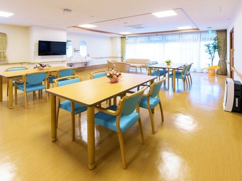 部屋の一面にはガラス窓が広がっており、縦型のブラインドカーテンが取り付けられている。ダイニングテーブルと青い椅子が置いてある。