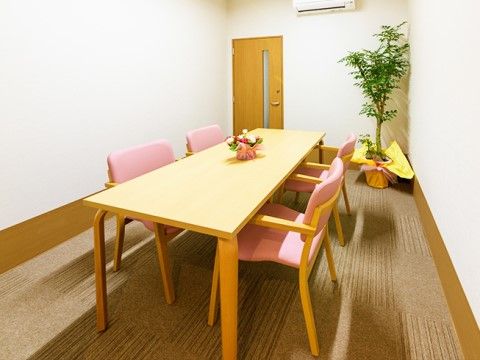 タイルカーペットが敷かれた部屋の角に、観葉植物が飾られている。部屋の中央にテーブルとピンク色の椅子が４脚置いてある。