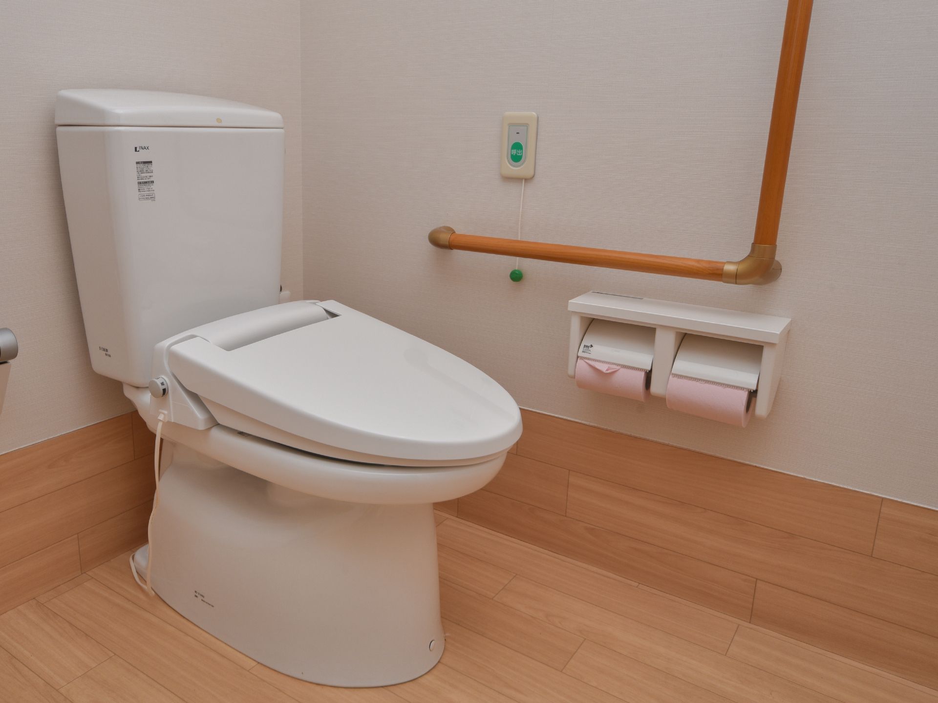 洋式トイレで、壁面に手すりが取り付けてある。トイレットペーパーのホルダーが二つ壁面についている。緊急時用のブザーが設置してある。