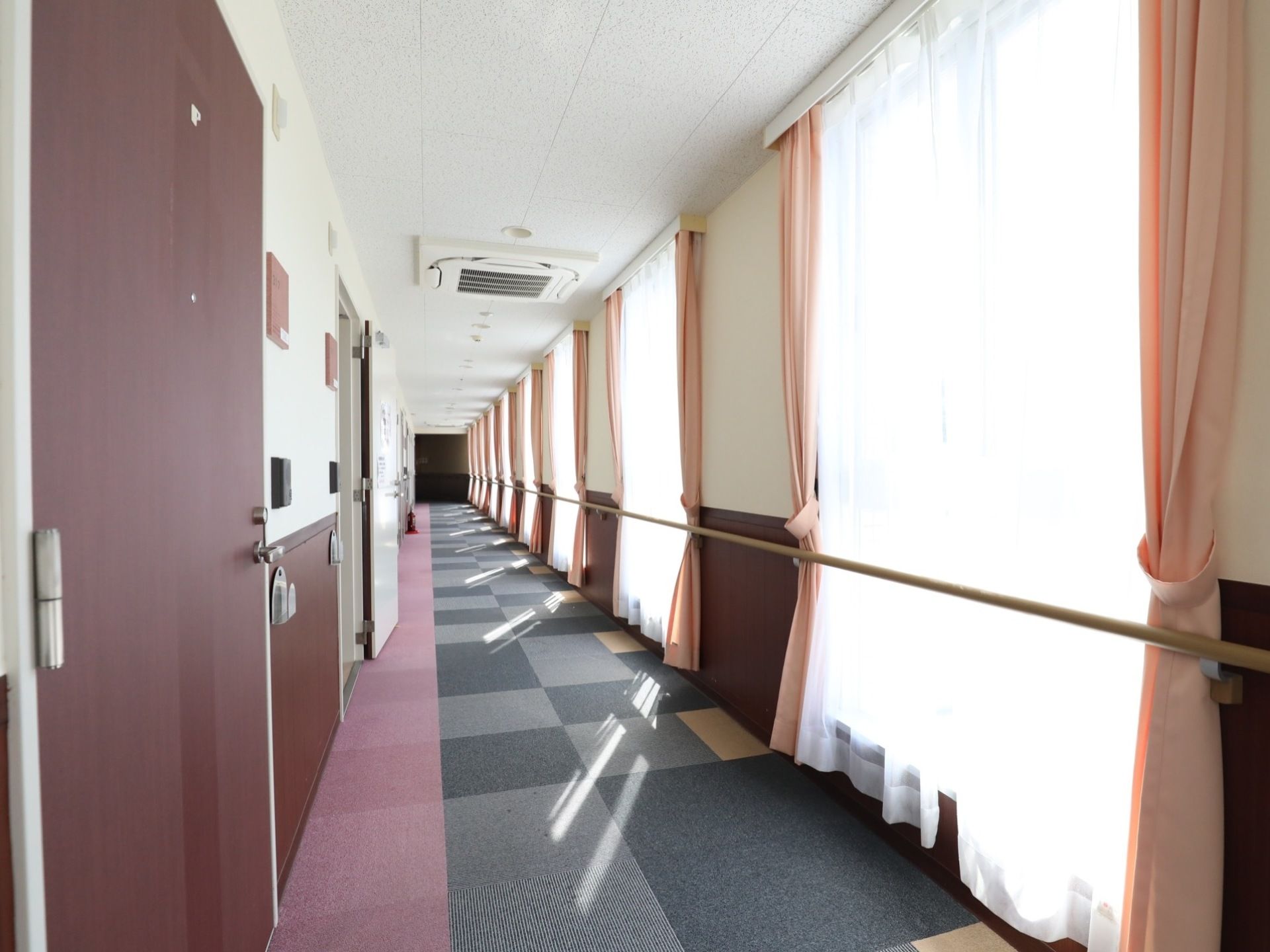 廊下はまっすぐに長く続いており、右側には大きな窓が続き、手すりが設置されている。左側には部屋のドアが並んでいる。