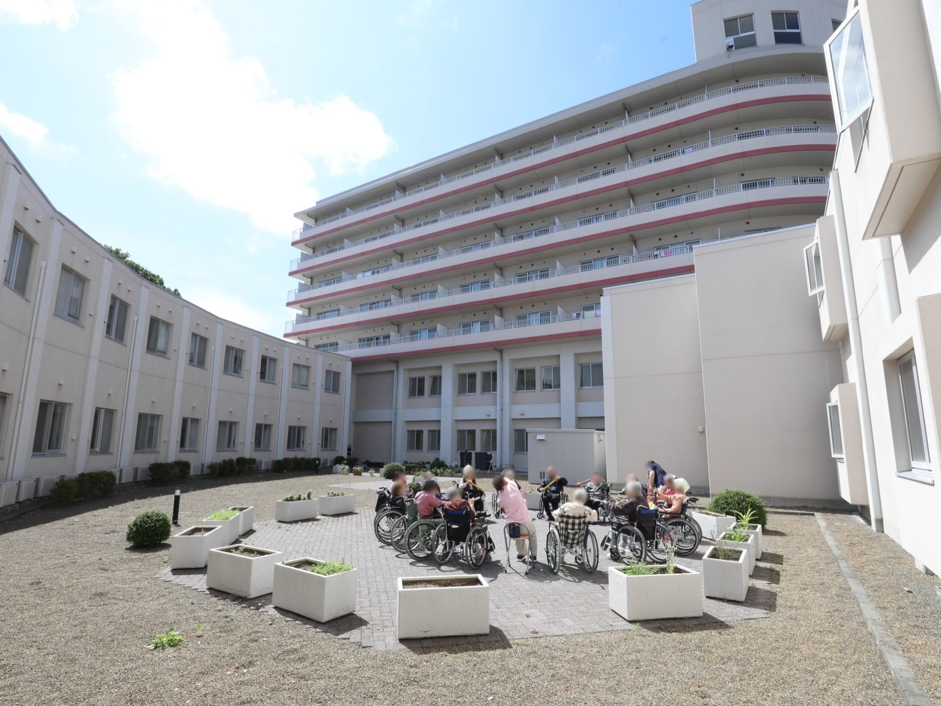施設の低い建物に囲まれた中庭は広く、中央にはプランターに囲まれたスペースがある。車椅子をご利用の方で輪になり体操をしている。