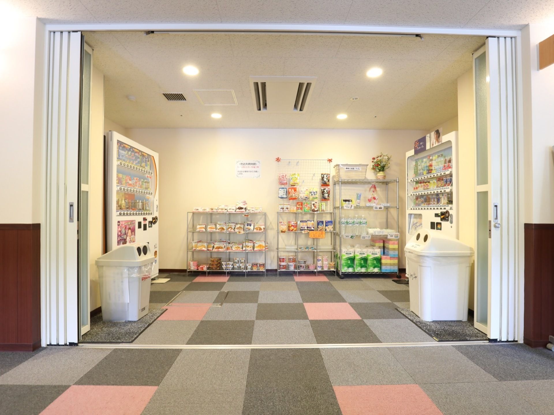 飲み物の自動販売機が２台、向かい合う形で設置されている。その間に棚があり、トイレットペーパーなどの日用品が並んで売られている。