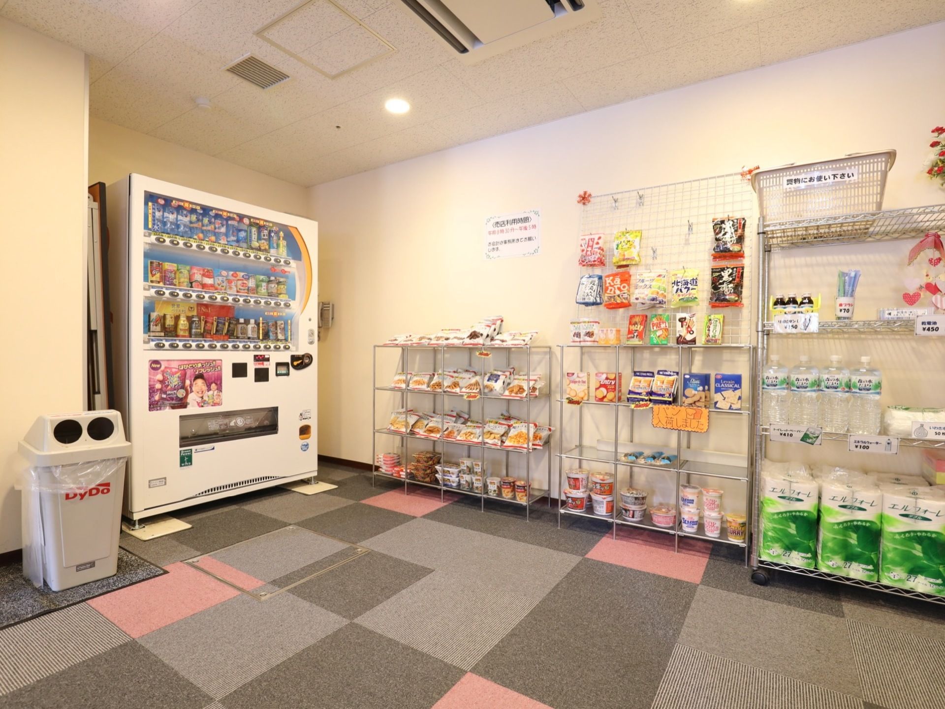 自動販売機の隣りにはごみ箱がある。売店内の棚には、パンやお菓子、水、トイレットペーパーなどが並んでいる。