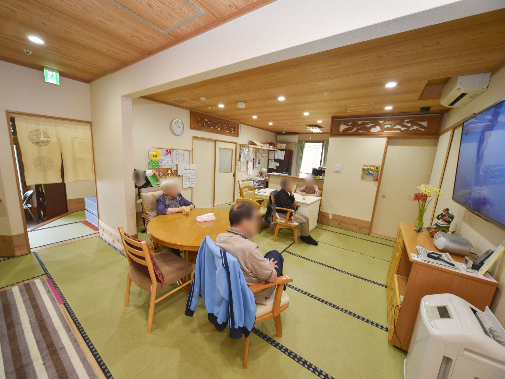 和室でくつろぐ利用者様の様子。右側にはテレビが設置されている。利用者様のそばで職員が仕事をし、一緒の空間で過ごしている。