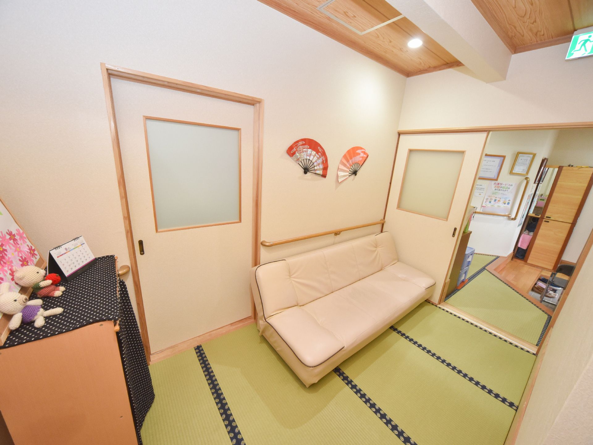 施設内の一室の様子。畳の部屋にソファが置かれている。壁には手すりがつけられている。壁面や棚の上には、扇子や人形が飾られている。