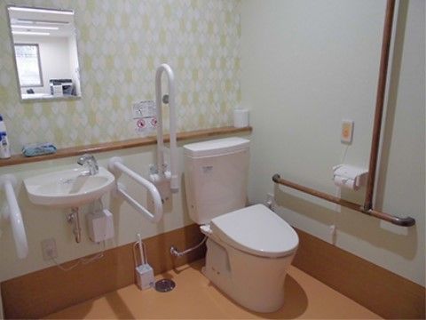 白い壁に薄いオレンジ色の床のトイレである。安全面と使いやすさを考慮した手すりがトイレの周りに設置されており、ナースコールも設置されている。