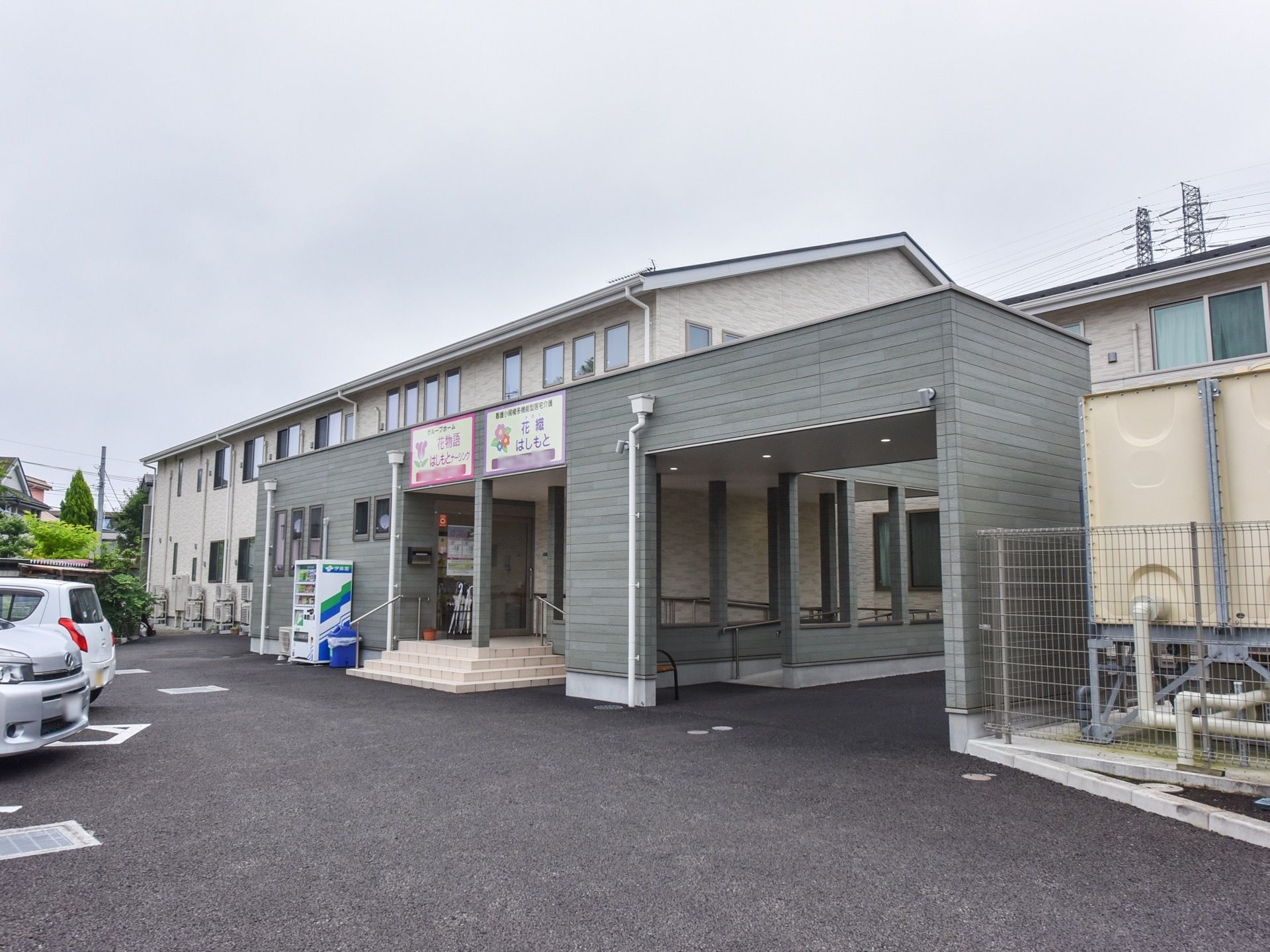 シックなグレーとベージュの2階建ての建物になっている。外には自動販売機も設置されている。駐車スペースも建物側に完備されている。