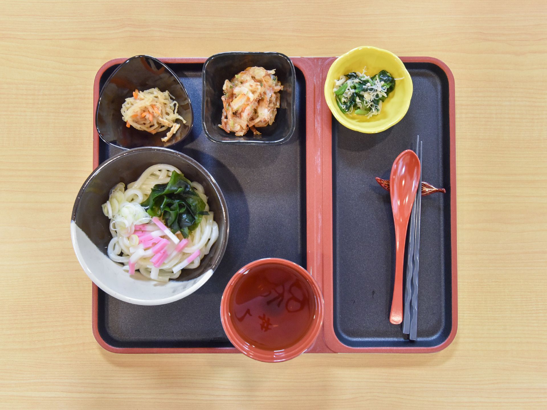 1人分の食事がセットされている様子である。箸は黒、樹脂製のスプーンは朱で、トレーはそれらのツートンカラーとなっている。