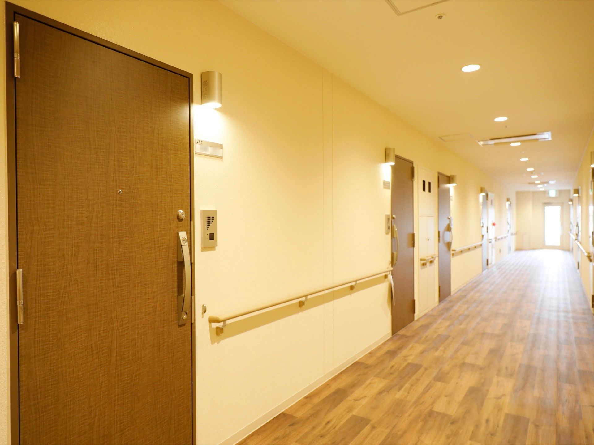 茶色の開き戸の居室のドアが並ぶ通路は、床面が淡い茶色の木目調で、壁はクリーム色で手すりが備わっている。