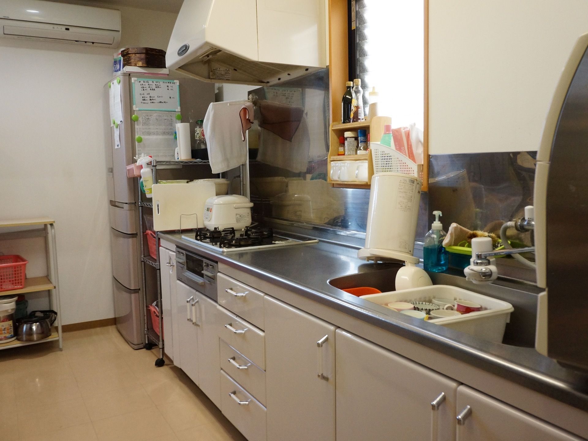 エアコンが完備されている。冷蔵庫、コンロ、キッチンシンクがある。洗い桶にはコップなどの食器が水に浸かっている。