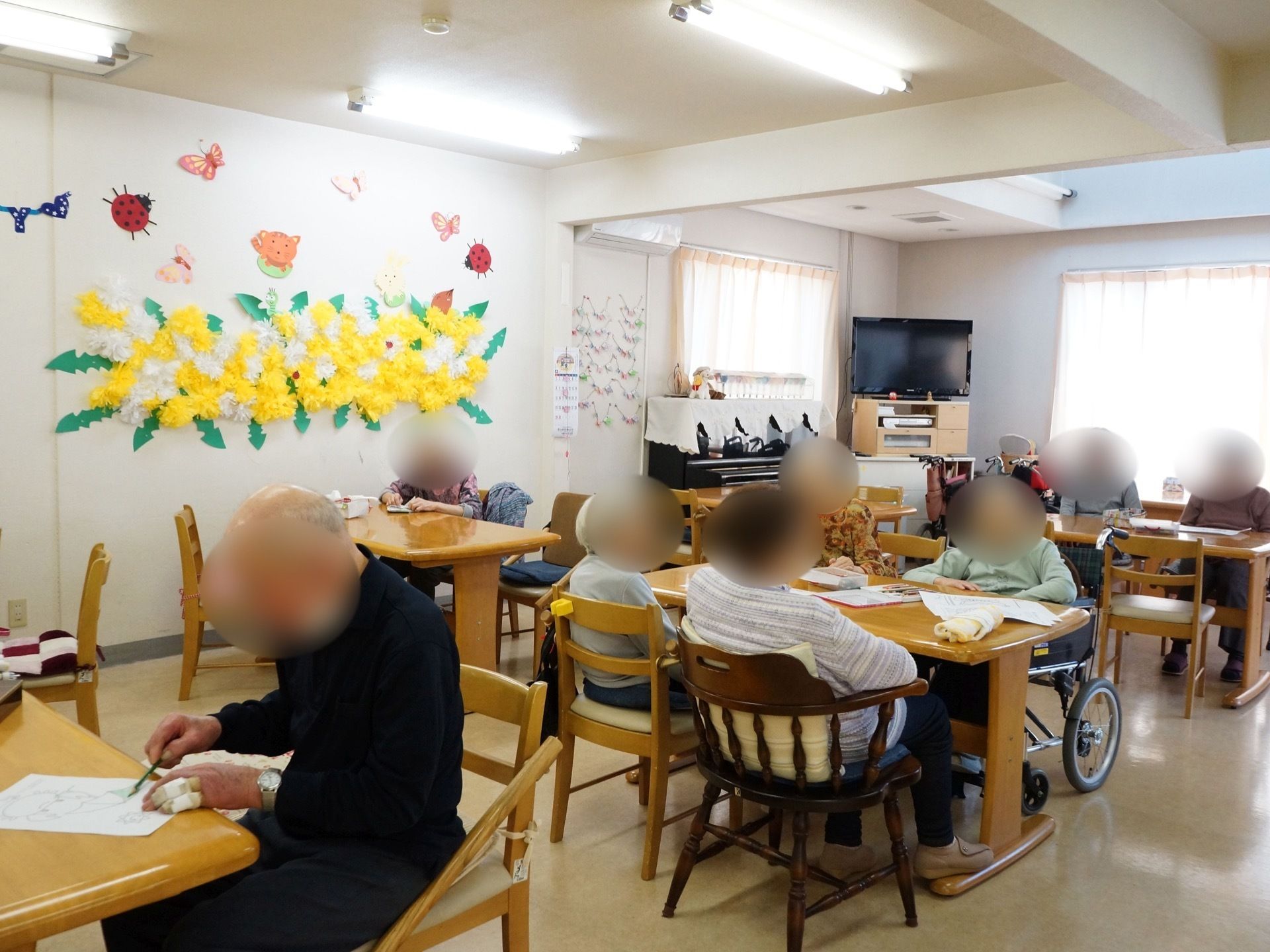 テーブルや椅子が並べられており、ご入居者様が作業をされている。ピアノが置かれている。壁には飾りつけがしてある。