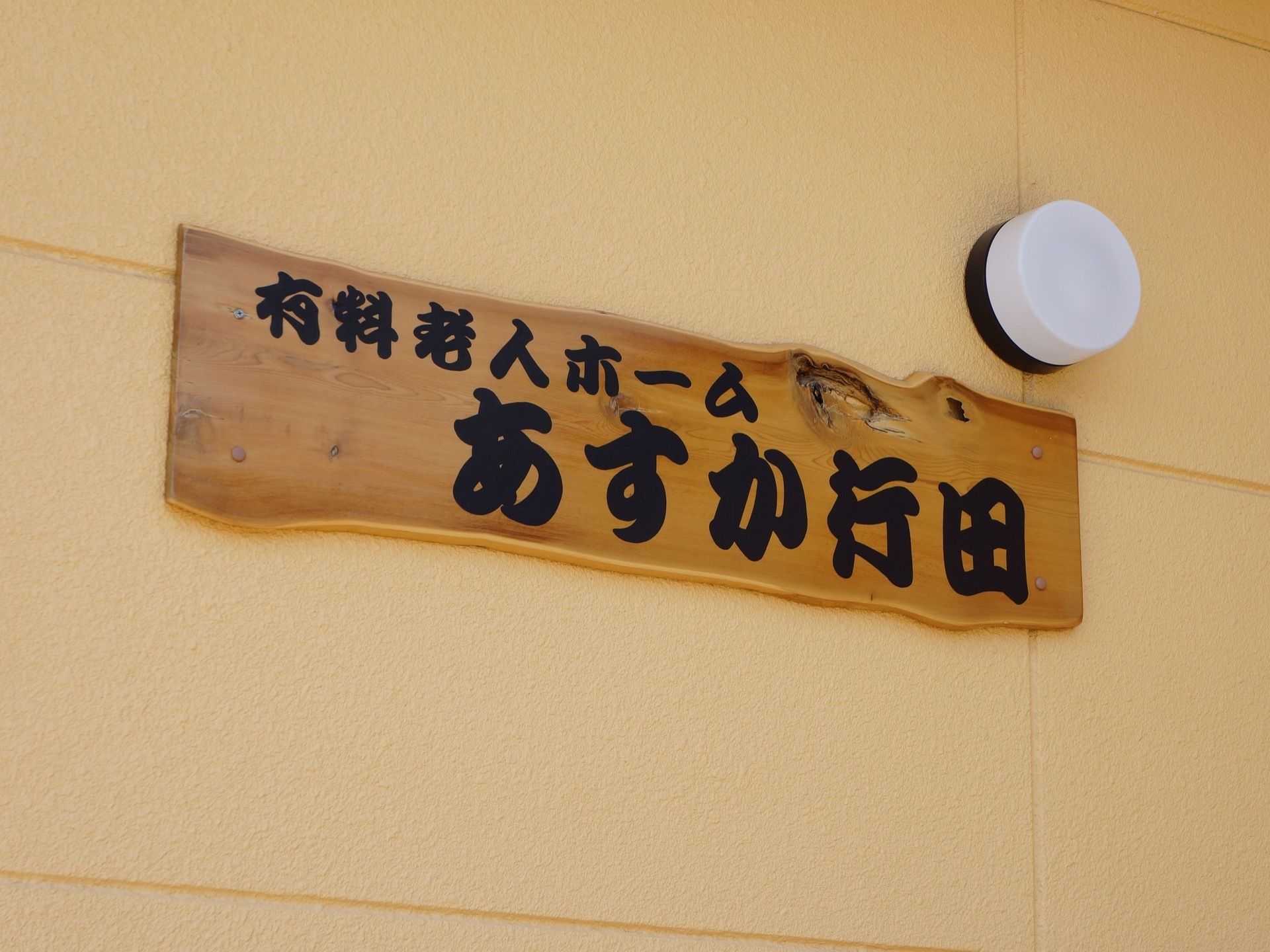施設名が力強い字体で大きく表示してある。外灯がある。看板は木目のある自然な木を使っている。外壁はアイボリー色である。