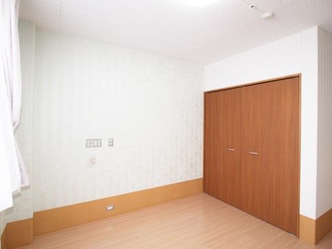 薄いストライプ模様の入った白いクロスの壁とフローリングの床のシンプルな部屋である。茶色い扉が取り付けられたクローゼットが設置されている。