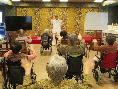 前に立つ男性が手の体操の見本を、それを真似する入所者たち