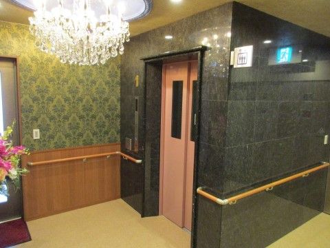 施設内のエレベーター。周囲の壁面はダークグリーンと落ち着いており、エレベーターの扉はピングと優しい彩りである。