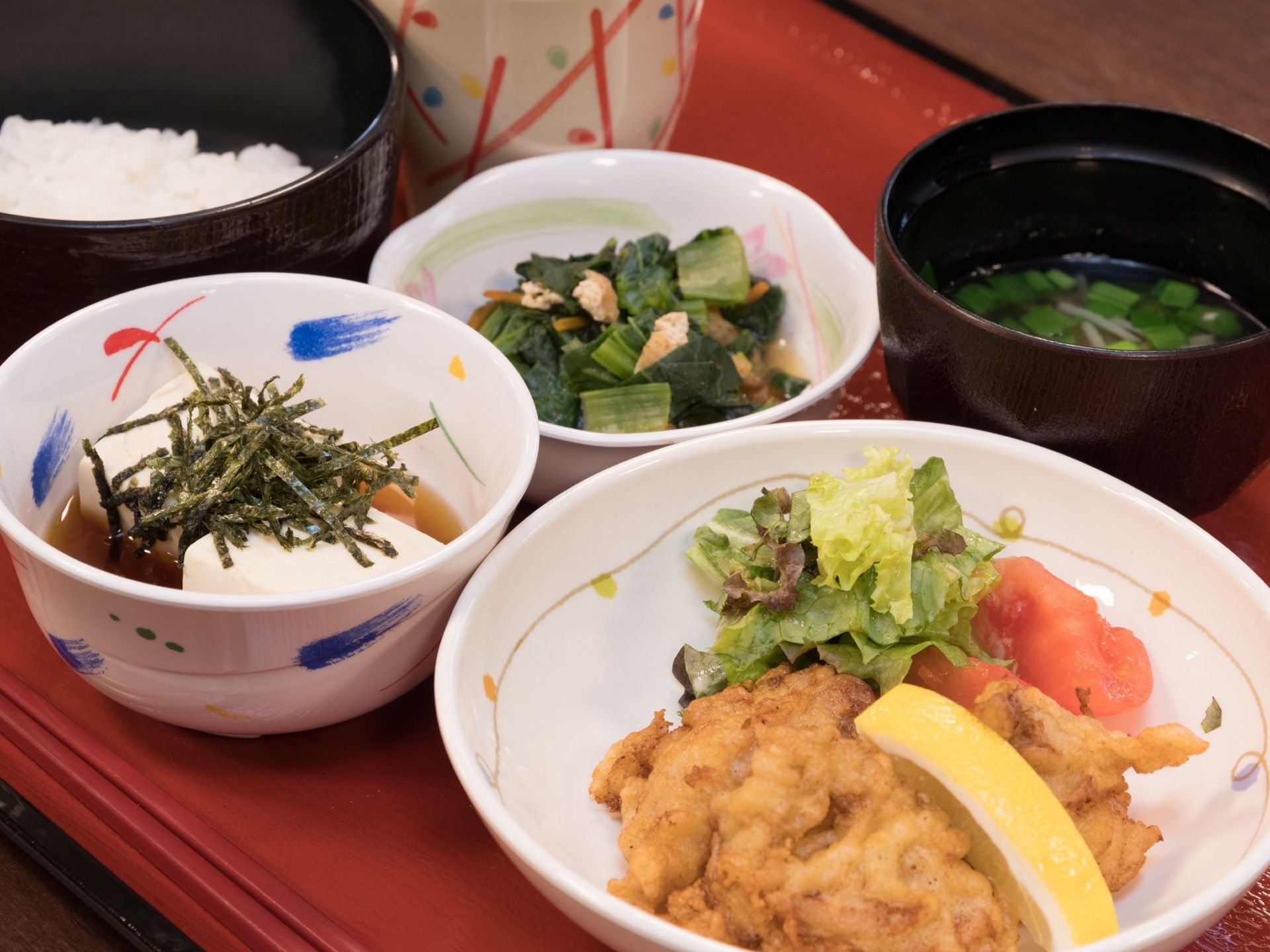 赤いお盆の上に食事が乗っている。メニューはご飯、味噌汁、冷奴、鳥のからあげ、ほうれん草の副菜とお茶である。
