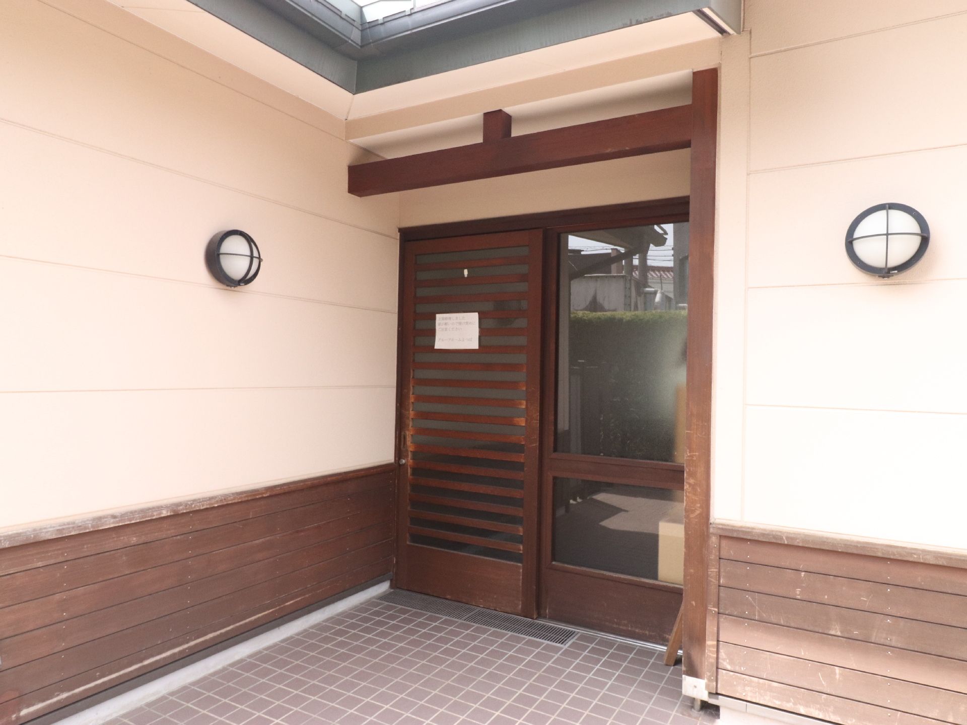 玄関扉は木製の引き戸になっている。化粧梁と外壁の一部にも木材が使われている。左右に和風の玄関灯が設置されている。