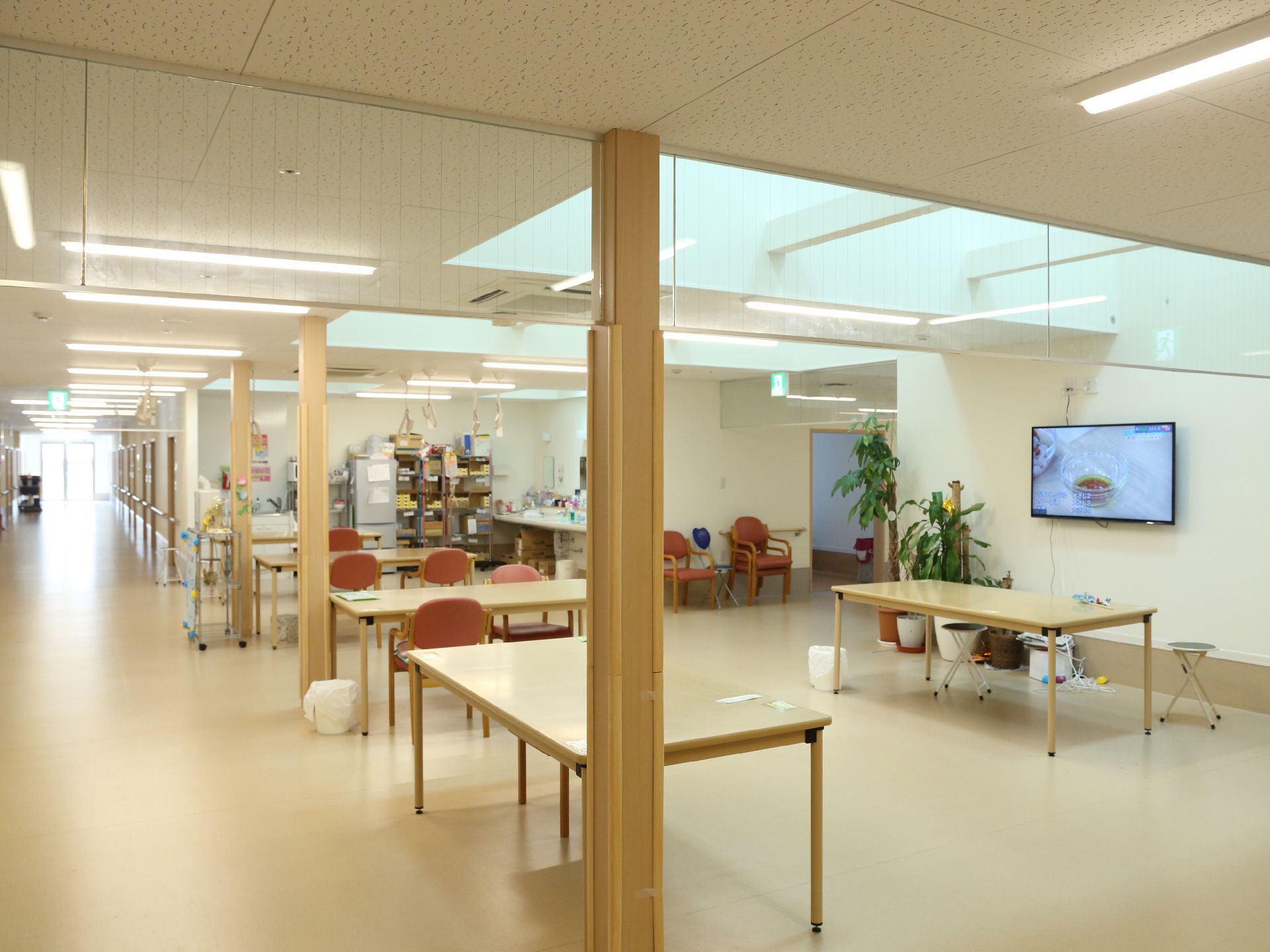 多くのテーブルと椅子が置かれている。右側の壁にはテレビが掛けられており、そばには観葉植物が置かれている。