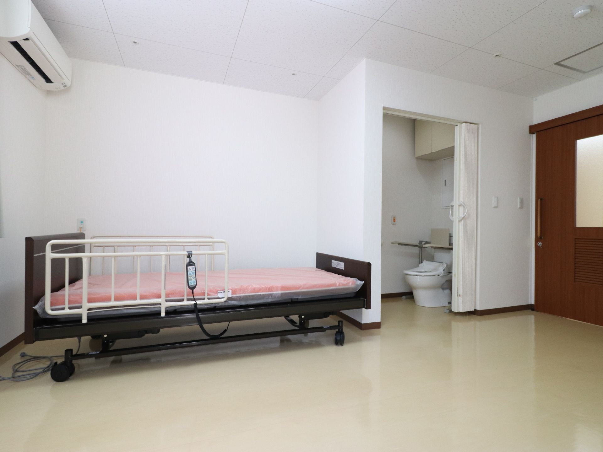 右側には扉がある。その左側にはトイレスペースがある。左側にはベッドがある。壁や天井は白色になっている。