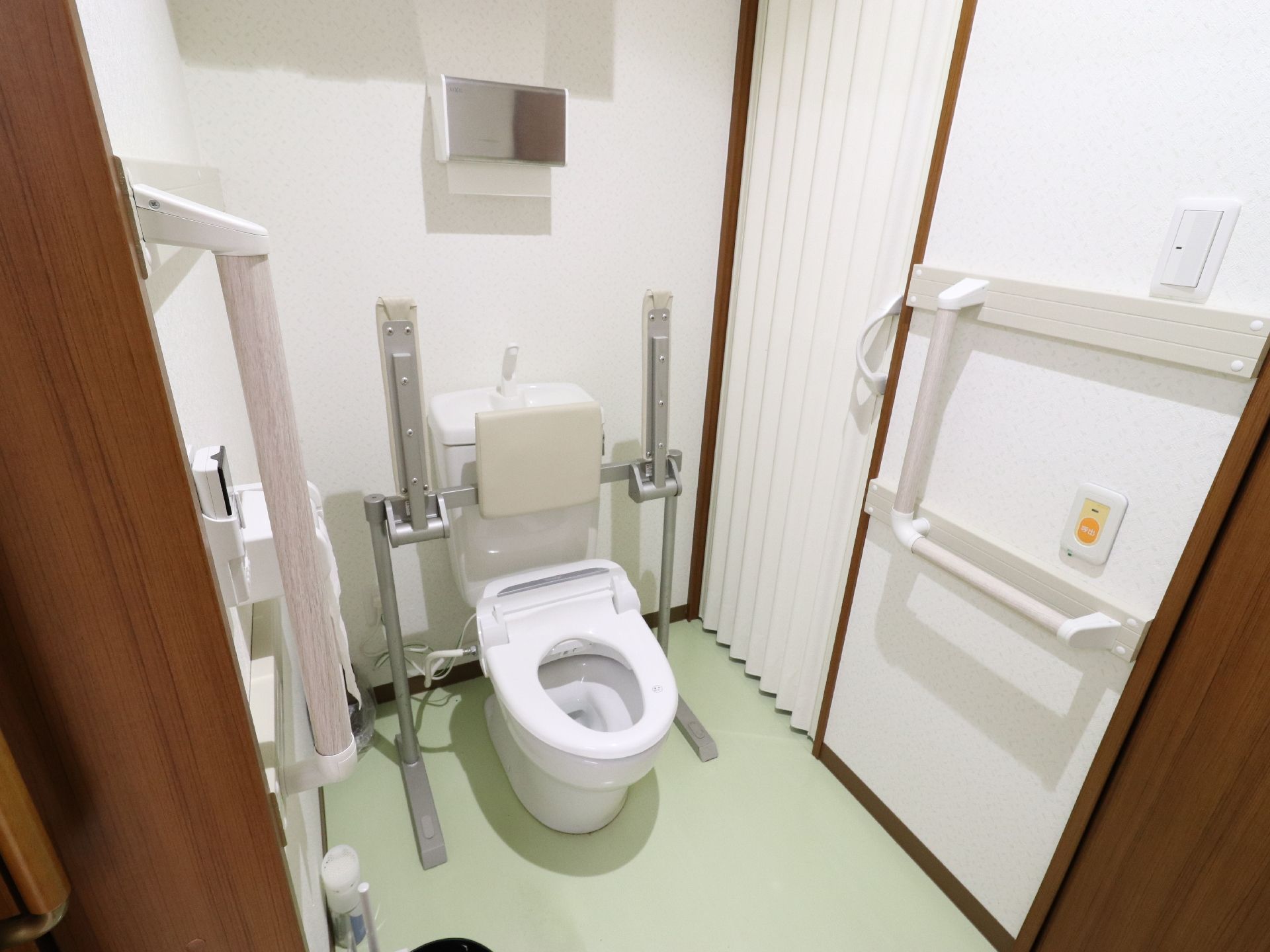 便器が正面にある。右側にはL字型の手すりが付いており、そばには緊急用の装置や電気のスイッチがある。奥にはペーパータオルがある。