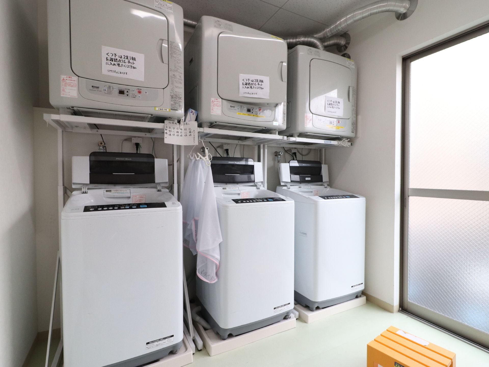 正面の下段には洗濯機が置かれている。上段には乾燥機があり、その取り出し口には張り紙が貼られている。それぞれ3台ずつ置かれている。