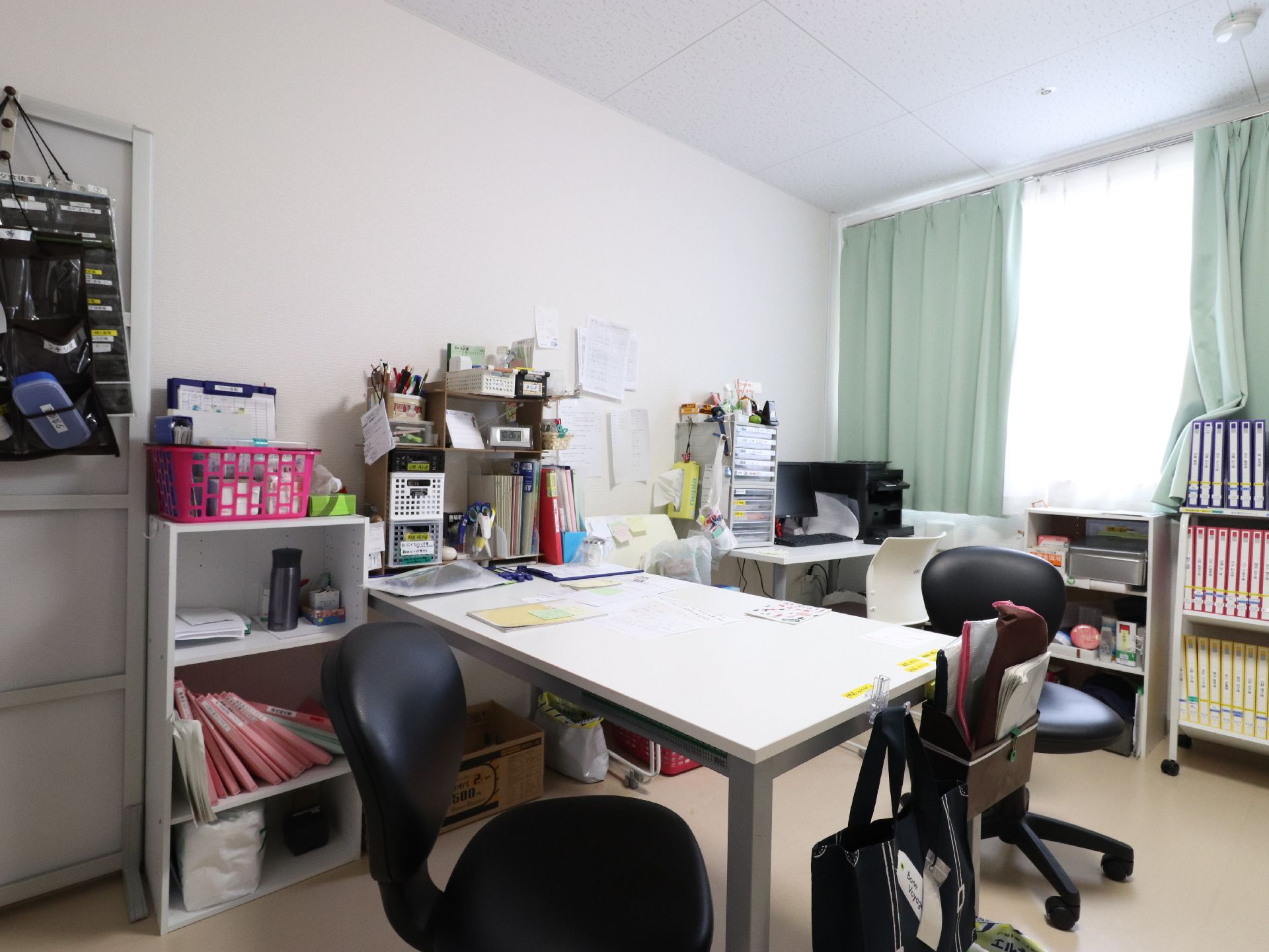 正面と奥にはテーブルがあり、そのそばには椅子が置かれている。右奥には収納棚があり、沢山のファイルが片付けられている。
