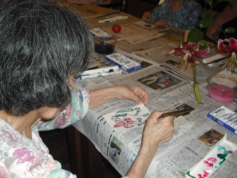新聞をひいた机の上で、百合を見ながら絵の具で絵手紙を描く女性