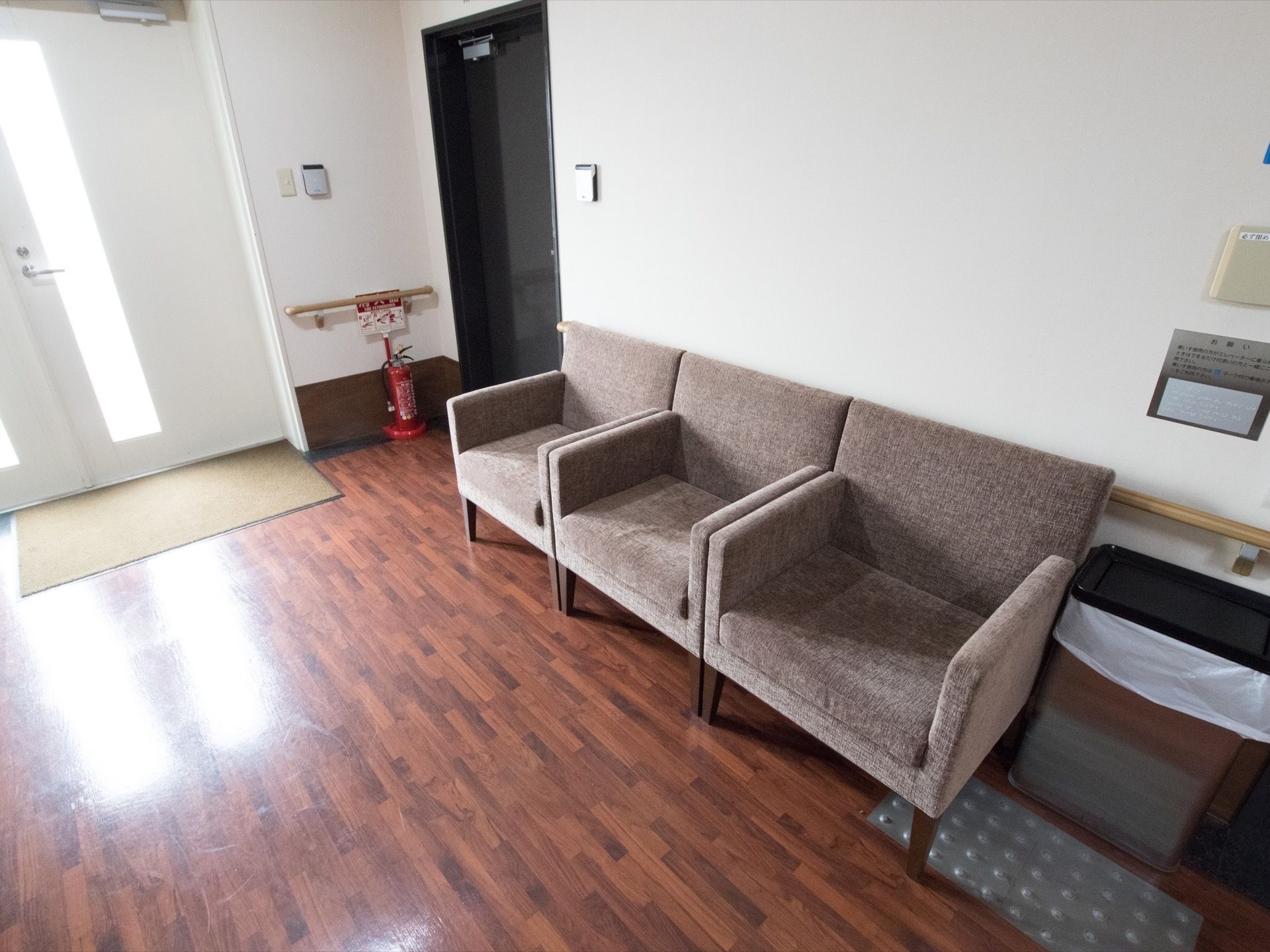 廊下の一角に１人掛けのソファが３脚置かれている。その横にゴミ箱が置かれている。奥に外へ出られる扉がある。