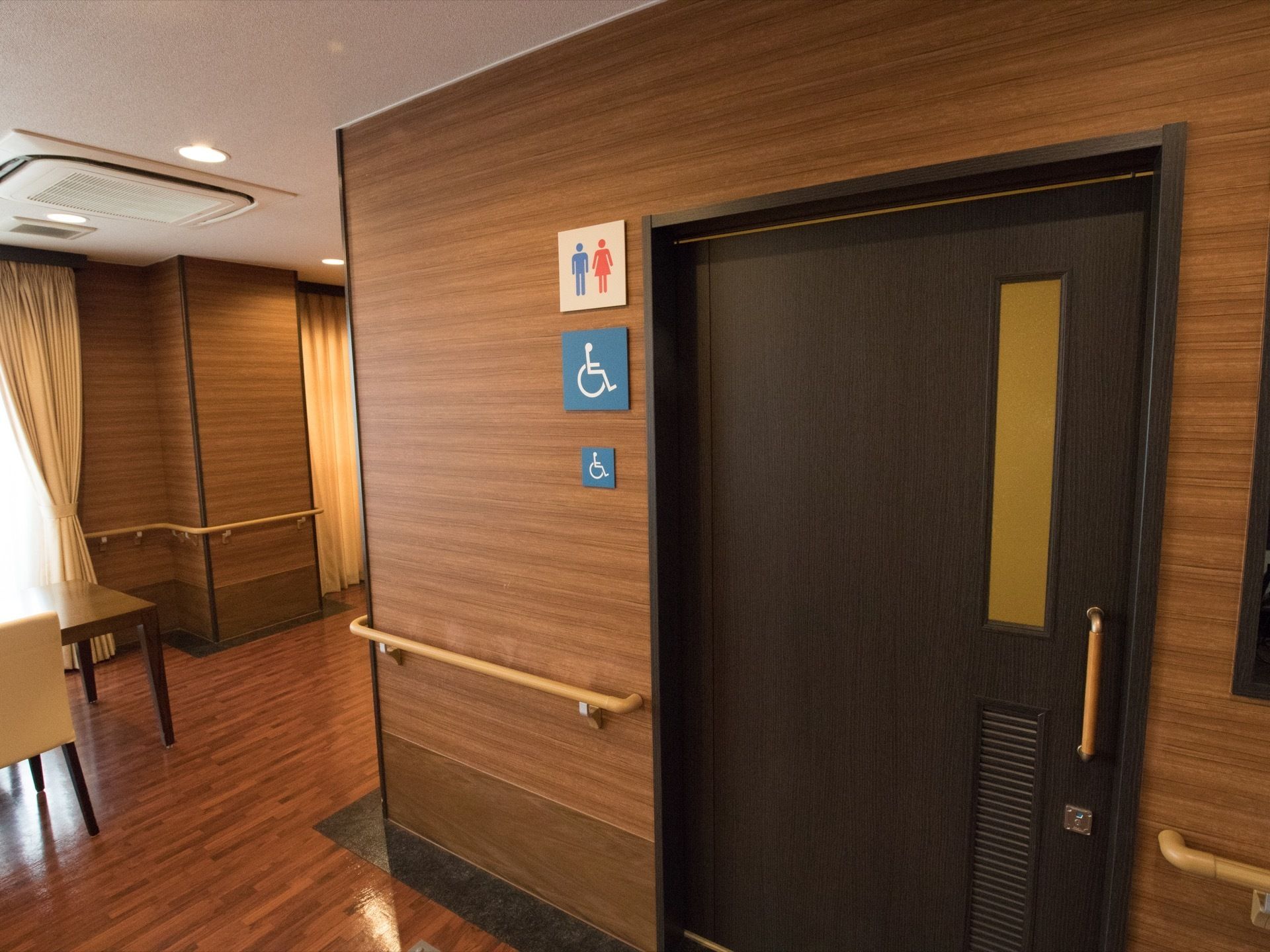 木目調の壁や扉で統一された廊下で、引き戸の扉の横にトイレのマークと車椅子用トイレのマークが貼られている。