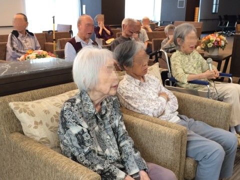 一人の車椅子女性がマイクを持ち、他の入居者も座ってくつろぐ様子