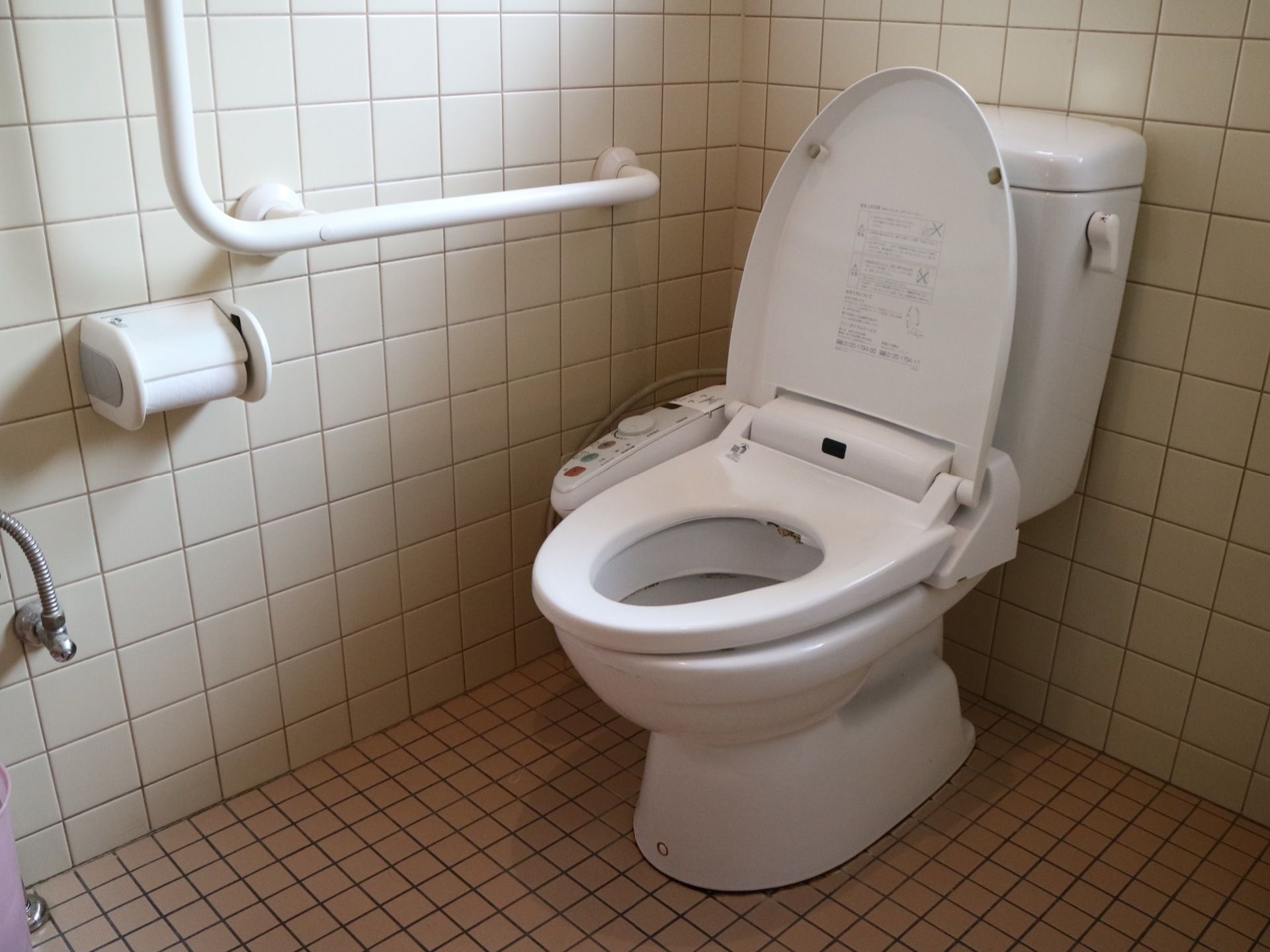 壁と床にタイルが貼られた個室トイレである。便座には温水洗浄機能が取り付けられている。壁に手すりとトイレットペーパーがある。