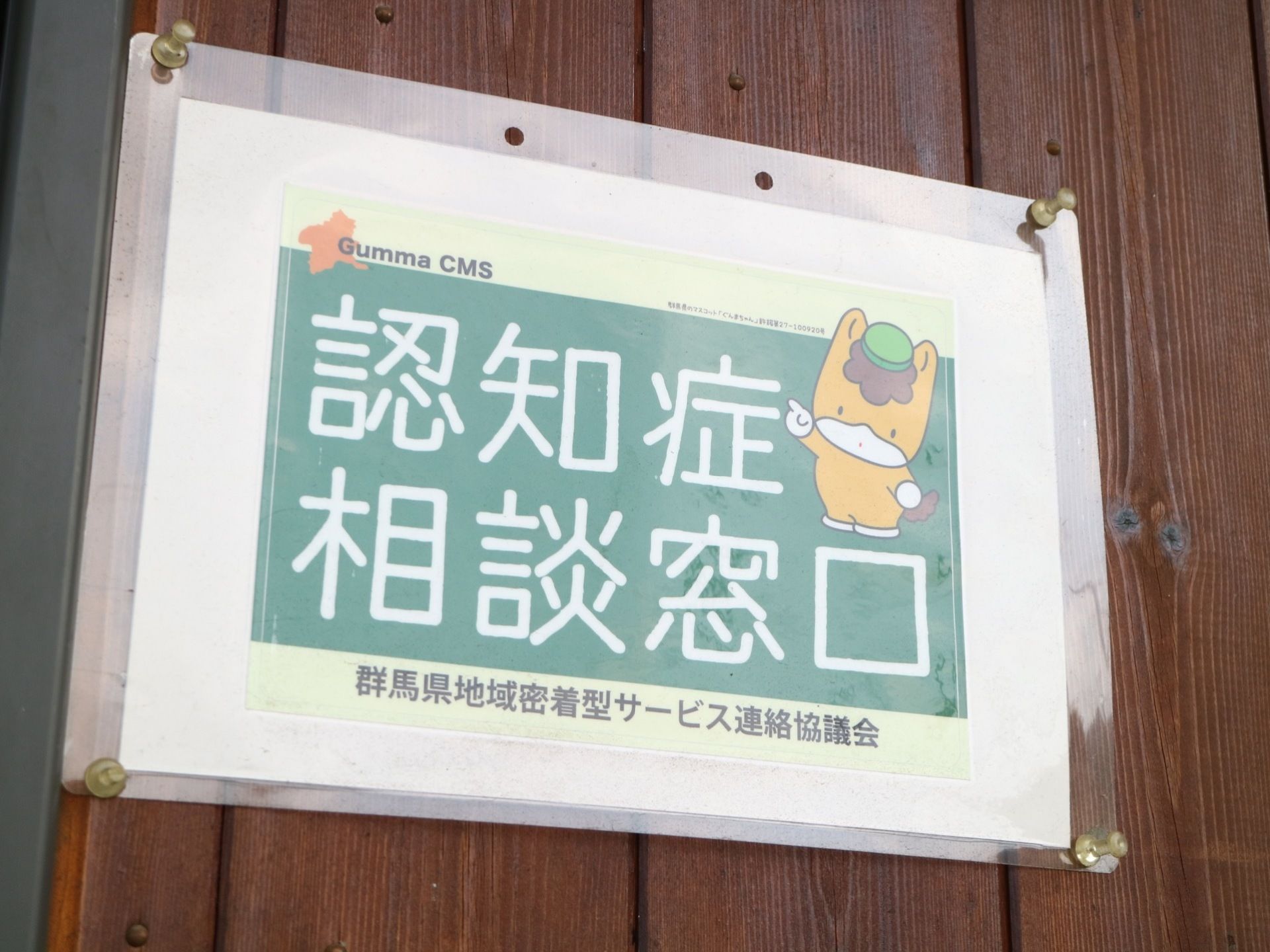 ダークブラウンの板材でできた壁に認知症相談窓口の貼り紙がされている。ご当地キャラクターのイラストが描かれている。