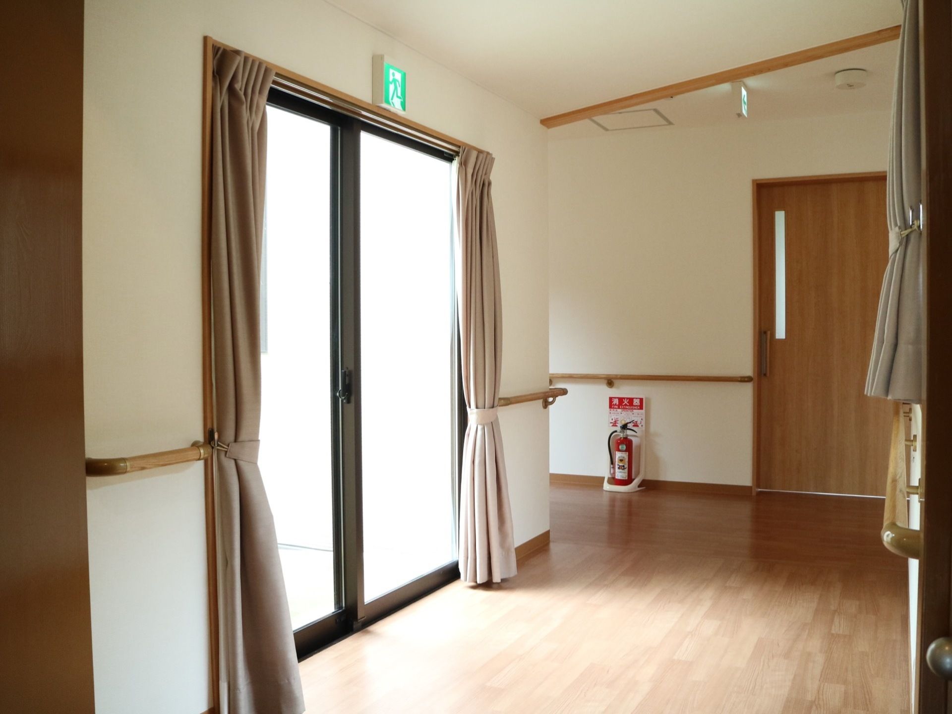 広々とした日当たりのよい廊下である。白を基調としているので清潔感がある。両側の壁には手すりが完備されている。消火器が置かれている。