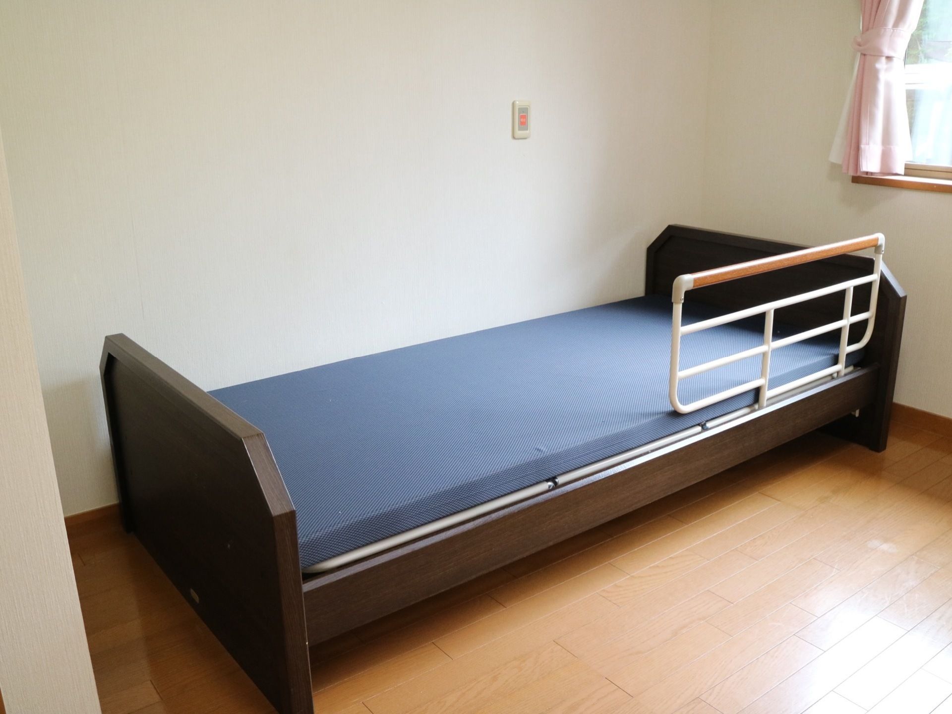 フローリングの居室に手すり付きのベッドが一台置かれている。ベッドの脇の壁には緊急用の通報装置が設置されている。