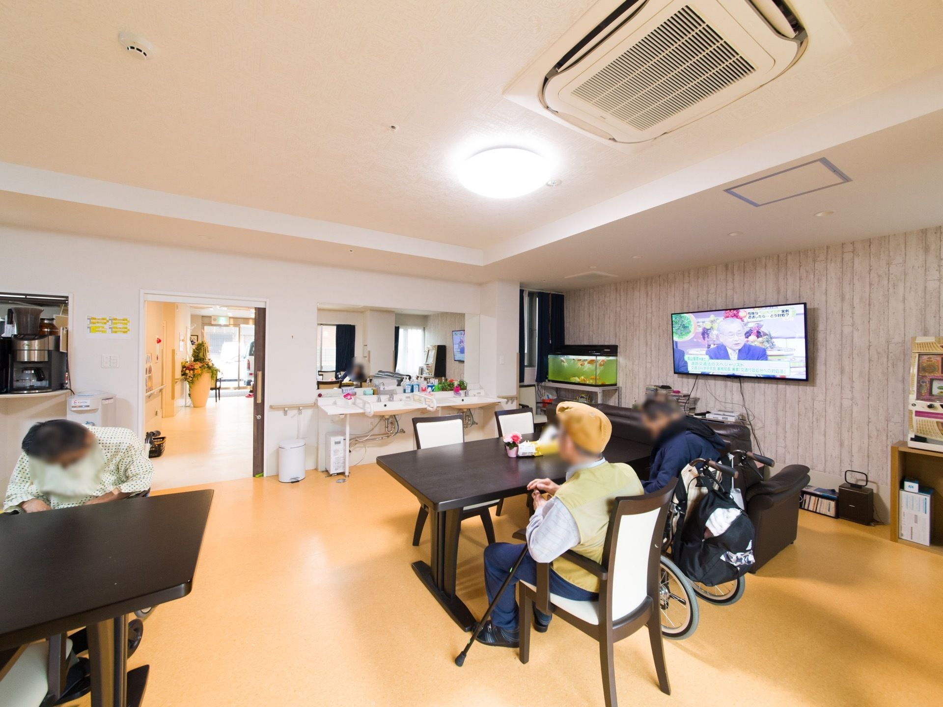 利用者様がくつろぐ共有スペース。テーブルと椅子が置かれ、テレビの前にはソファーがある。入り口の右わきに洗面台があり、左側にはカウンターがある。
