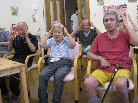 椅子に座った数名の利用者様が楽しそうに頭に手を当てている様子